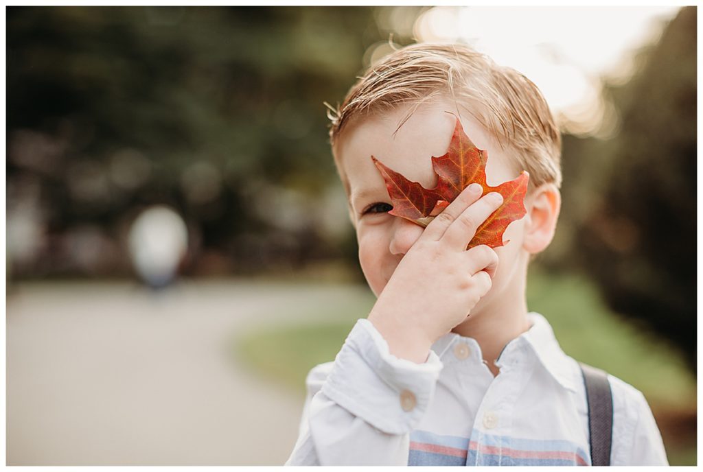 little boy in suspenders holding a red autumn leaf in front of his eye