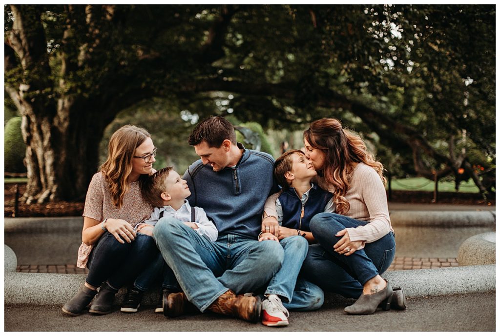 Family of five in peach and navy blue outfits sitting on the edge of a fountain and kissing and laughing. 