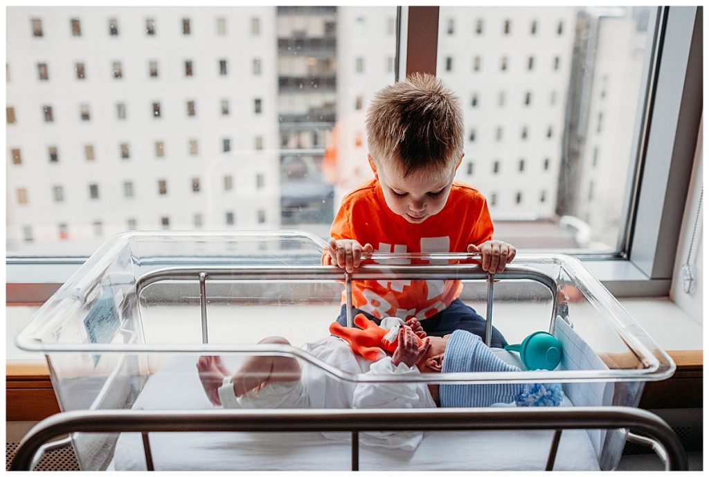 toddler-boy-peeking-over-side-of-hospital-bassinet-boston-newborn-photographer