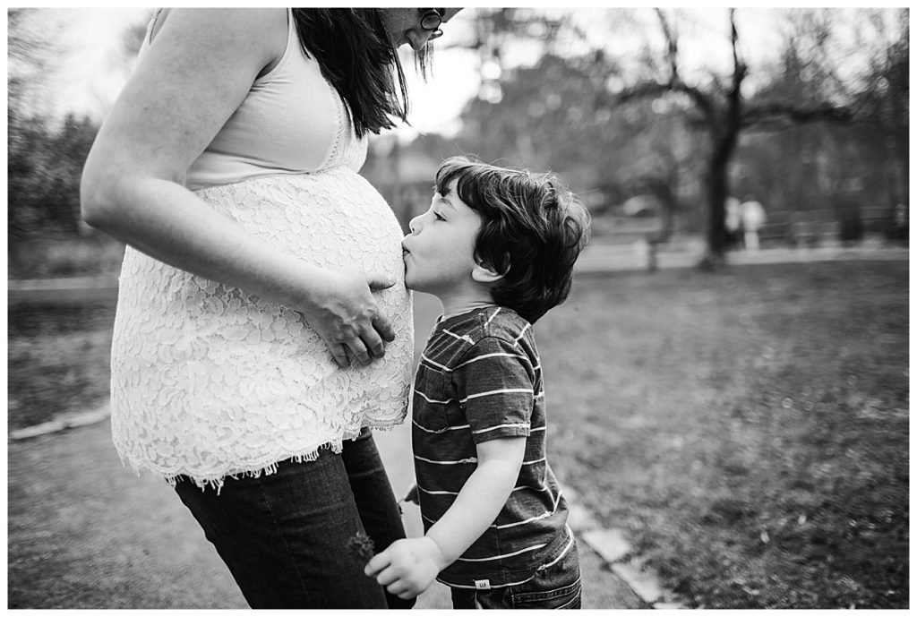 black and white portrait of young boy kissing pregnant mother's baby bump during family photos