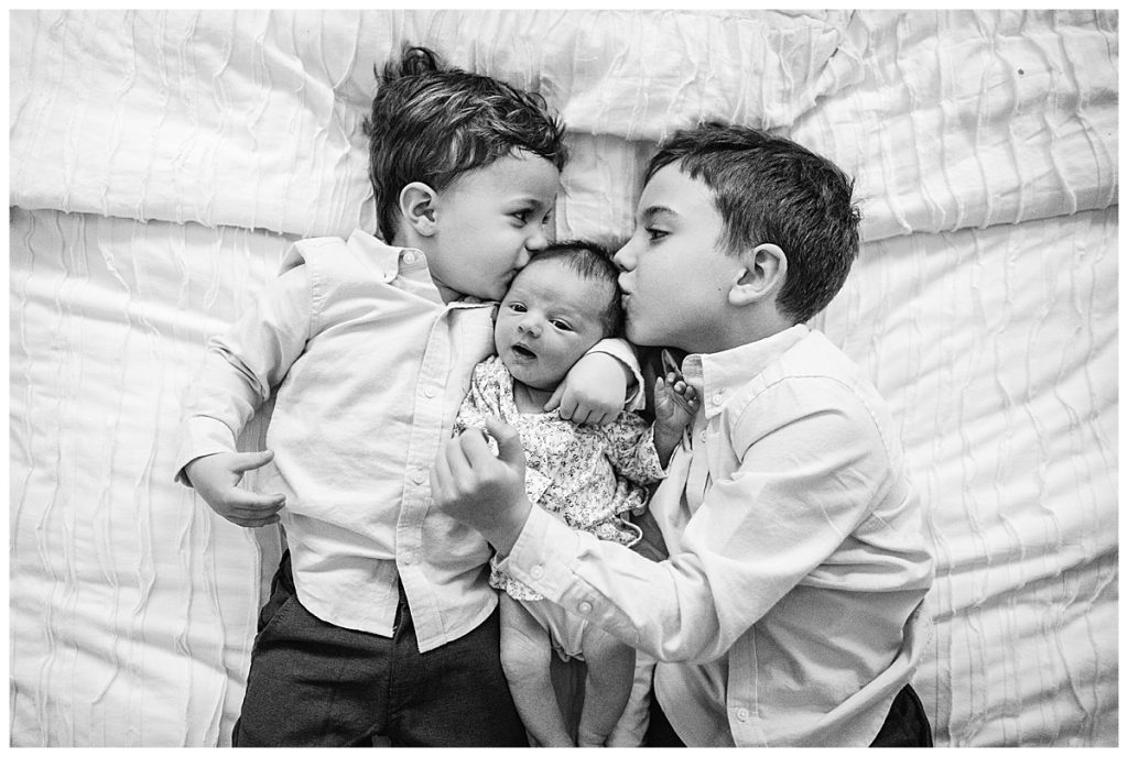 black and white image of big brothers kissing baby sister during newborn pics