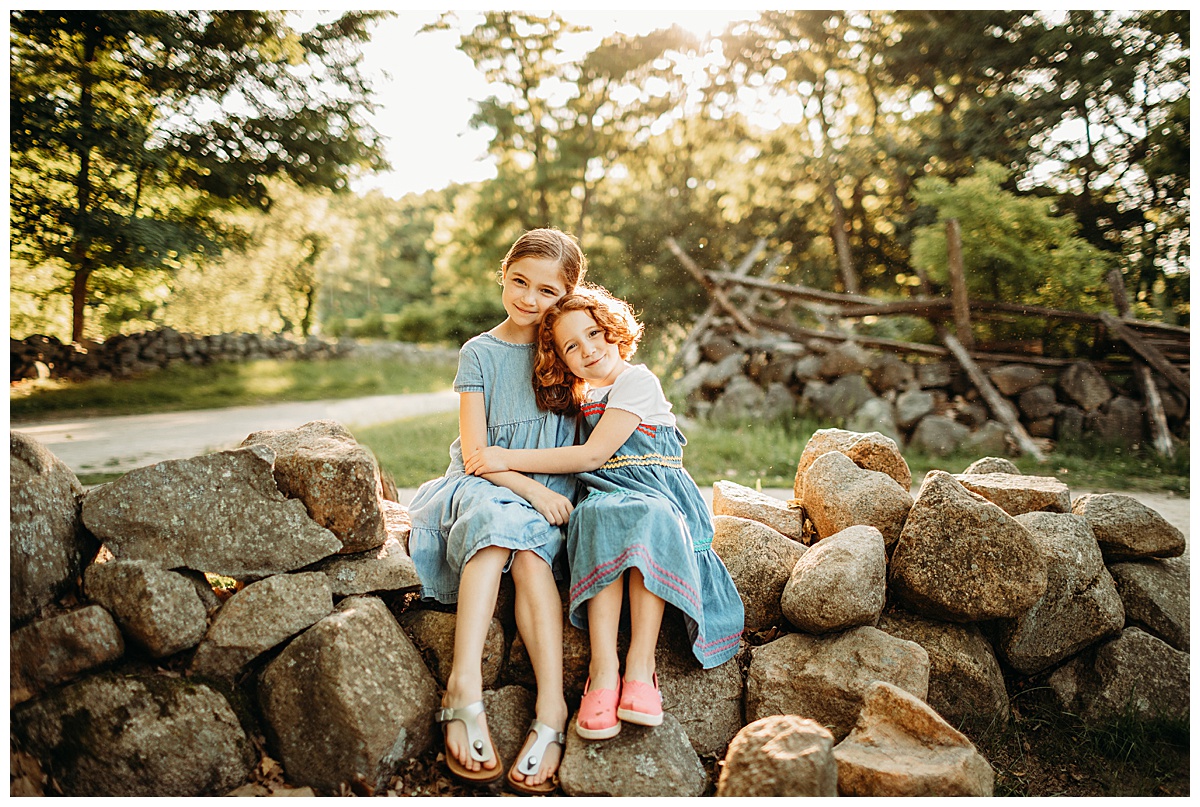 sisters in denim dresses sit on a stone wall during family photo session