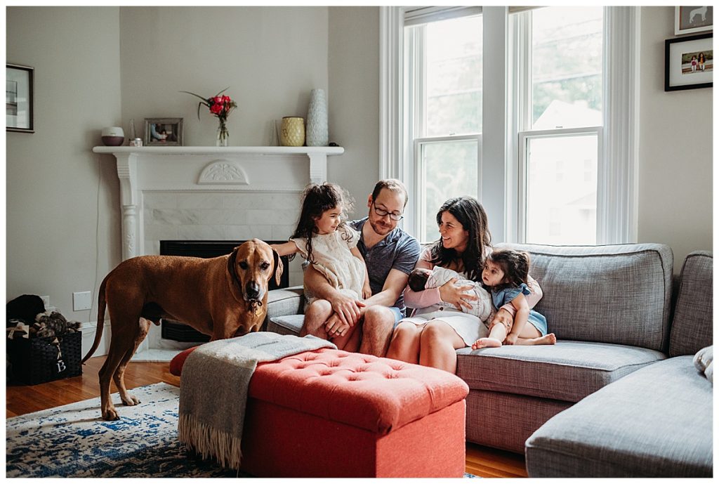 family sits on couch in living room with new baby daughter and large dog