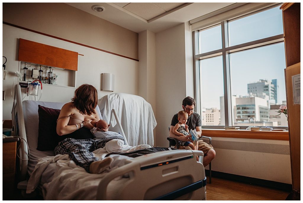 mother feeds newborn baby in hospital bed while dad reads to sister