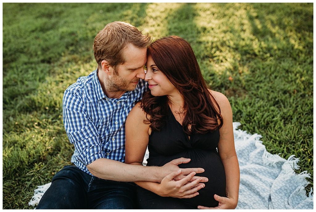 pregnant couple sit on blanket outdoors and hold baby bump