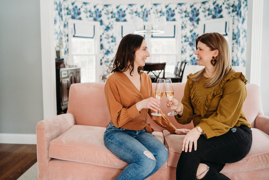 two women clink champagne glasses on a pink couch during their personal brand photography session in sudbury