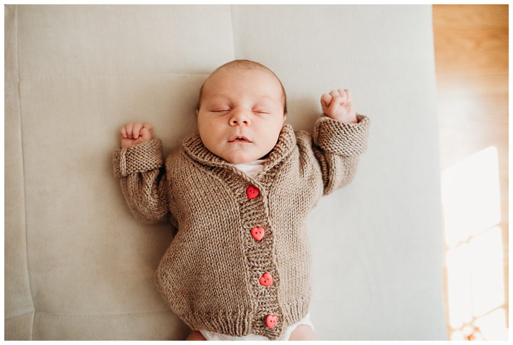 newborn is asleep in brown sweater with pink buttons during photography at home in holliston ma 