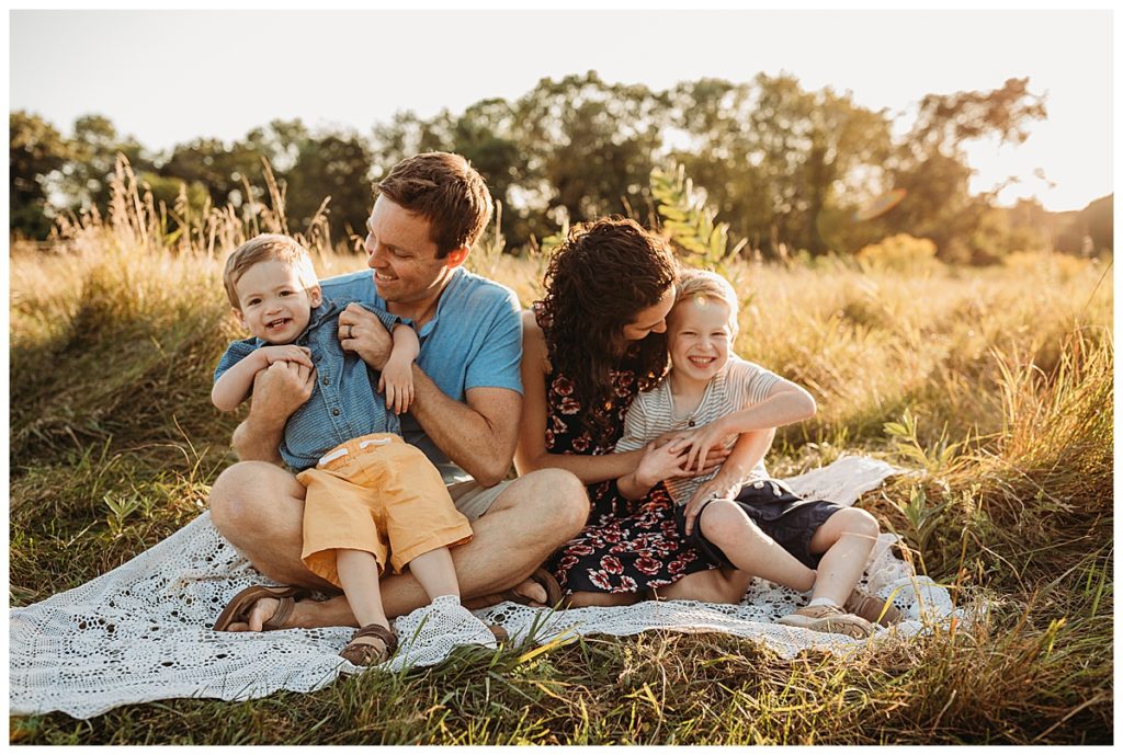 family sits on lace blanket outdoor in a field at sunset