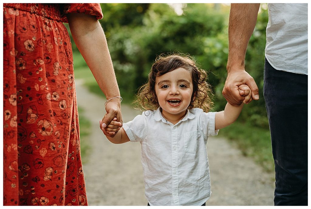 toddler grins at camera while holding parents hands