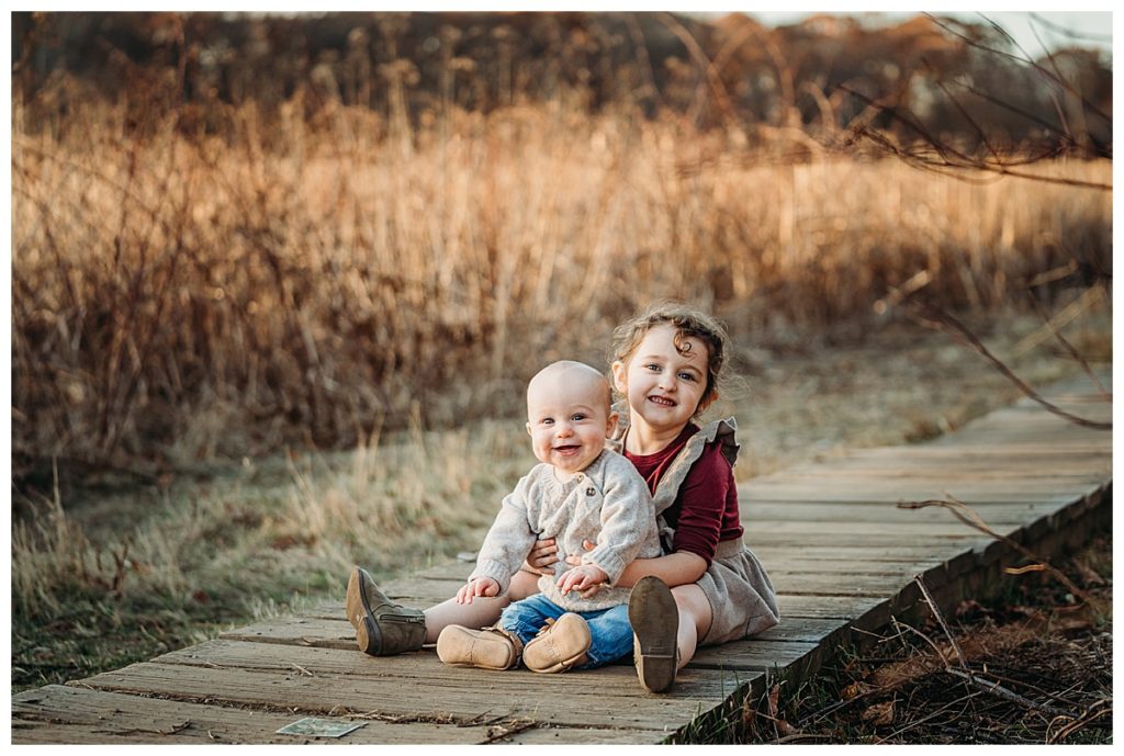 siblings sit on wooden path together and smile at the camera
