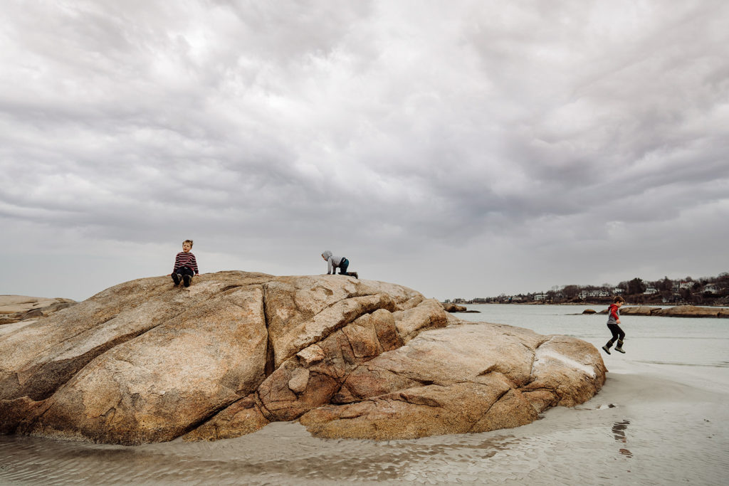 three boys climb huge rocks at the beach and jump off of them