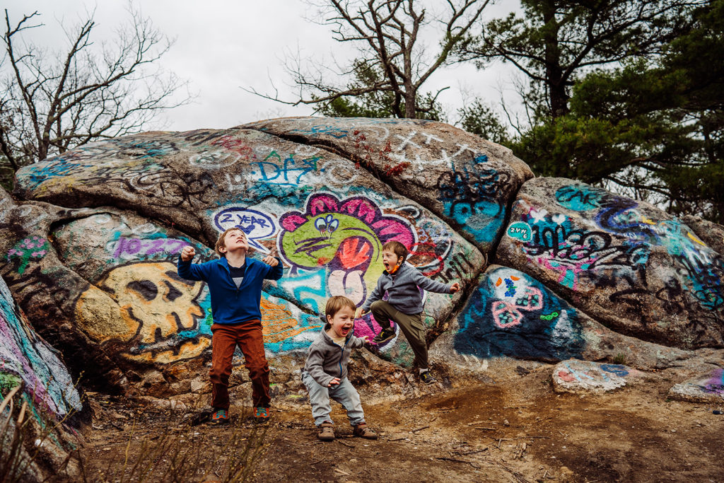 three boys in hiking clothes jump and scream in front of a graffiti covered rocky cliff