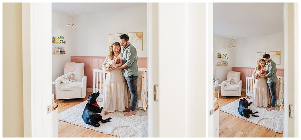 parents with new baby girl stand inside pink nursery while black lab looks up at them
