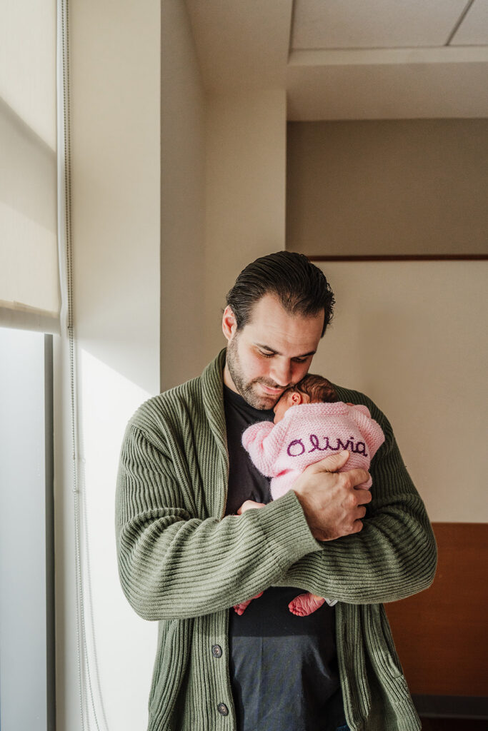 a dad in a green cardigan snuggles new baby girl wearing a pink sweater next to boston hospital window