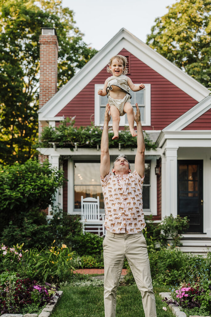 Candid family photo near Boston with dad tossing toddler, coordinated summer outfits.